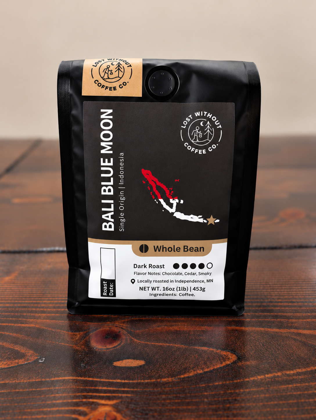 A bag of Bali Blue Moon whole bean coffee sits on a dark wooden tabletop. The packaging is matte black with a tan label at the top and a large tan, white, and red front label.